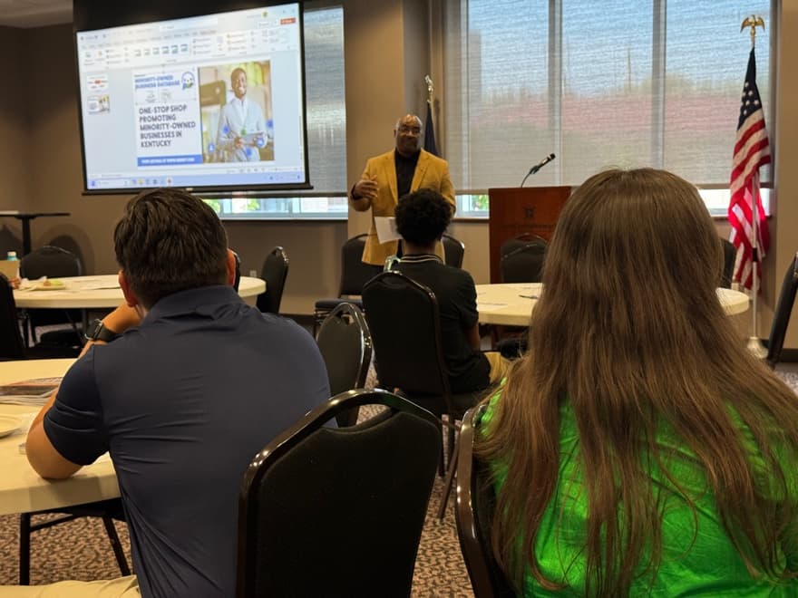 A presenter in a gold jacket speaking beside a projected slide about a minority-owned business database, seen from behind seated attendees in a conference room with an American flag