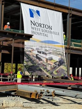 A Norton West Louisville Hospital banner hung from the steel frame of the hospital under construction, with workers in safety vests on the deck around it