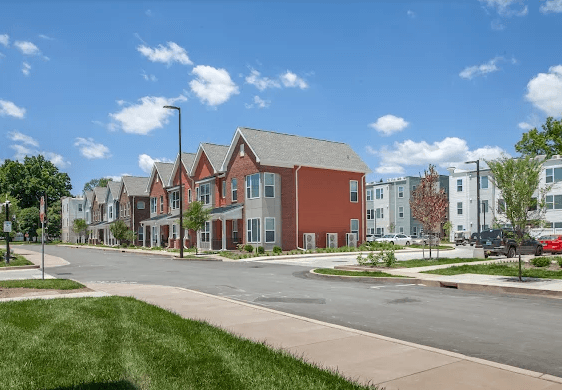 A newly built street of brick and grey townhomes with young trees and fresh sidewalks under a blue sky